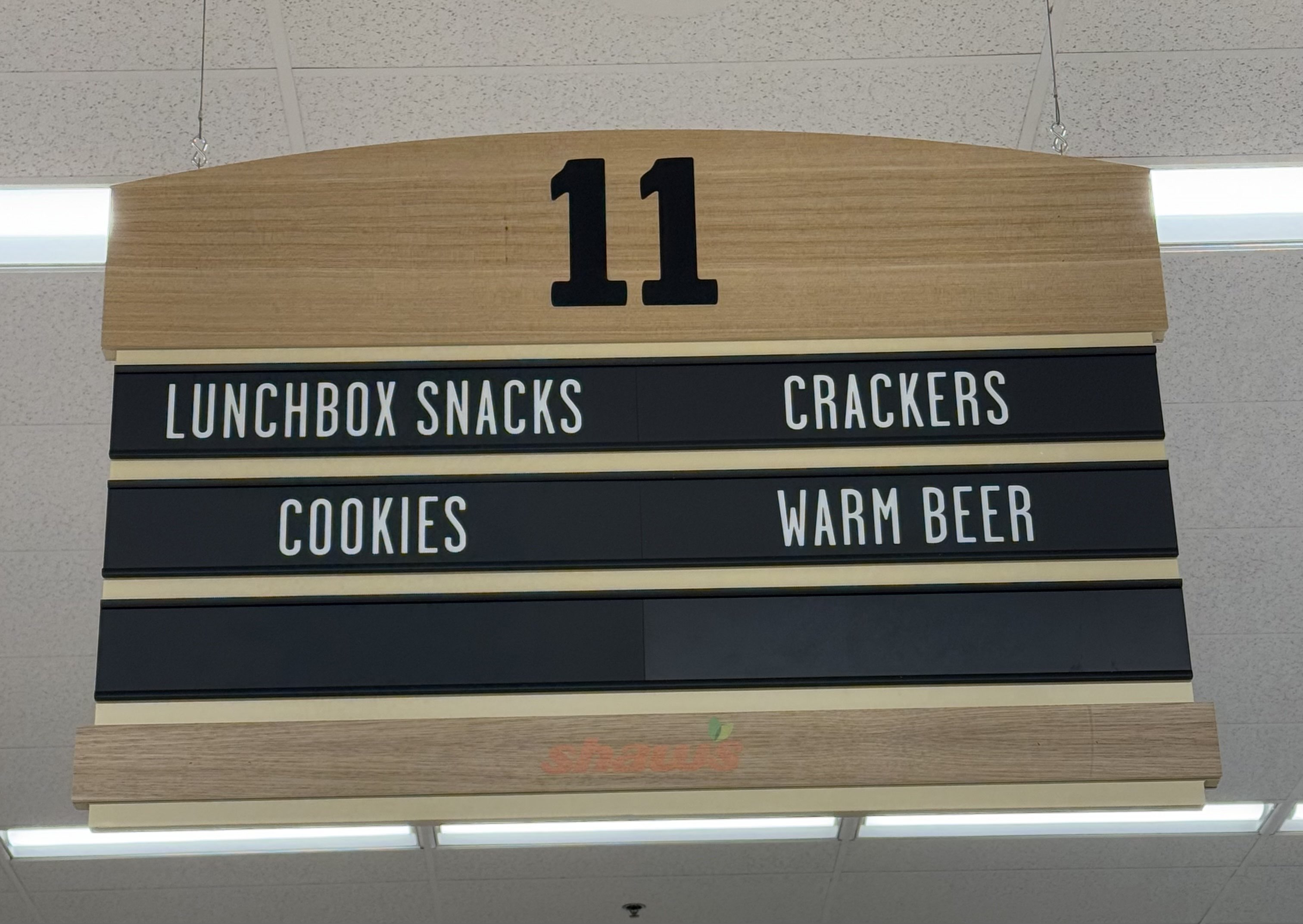 A grocery store aisle sign advertising lunchbox snacks, crackers, cookiers, and warm beer.
