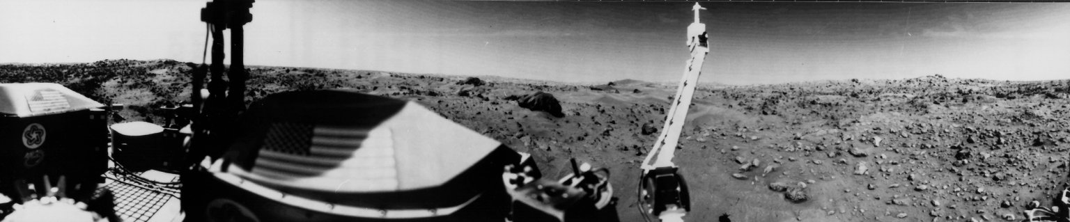 A black and white panoramic photo of the surface of Mars, taken by the Viking 1. The surface of the planet is rocky and flat. The Viking 1 itself takes up a large portion of the frame.